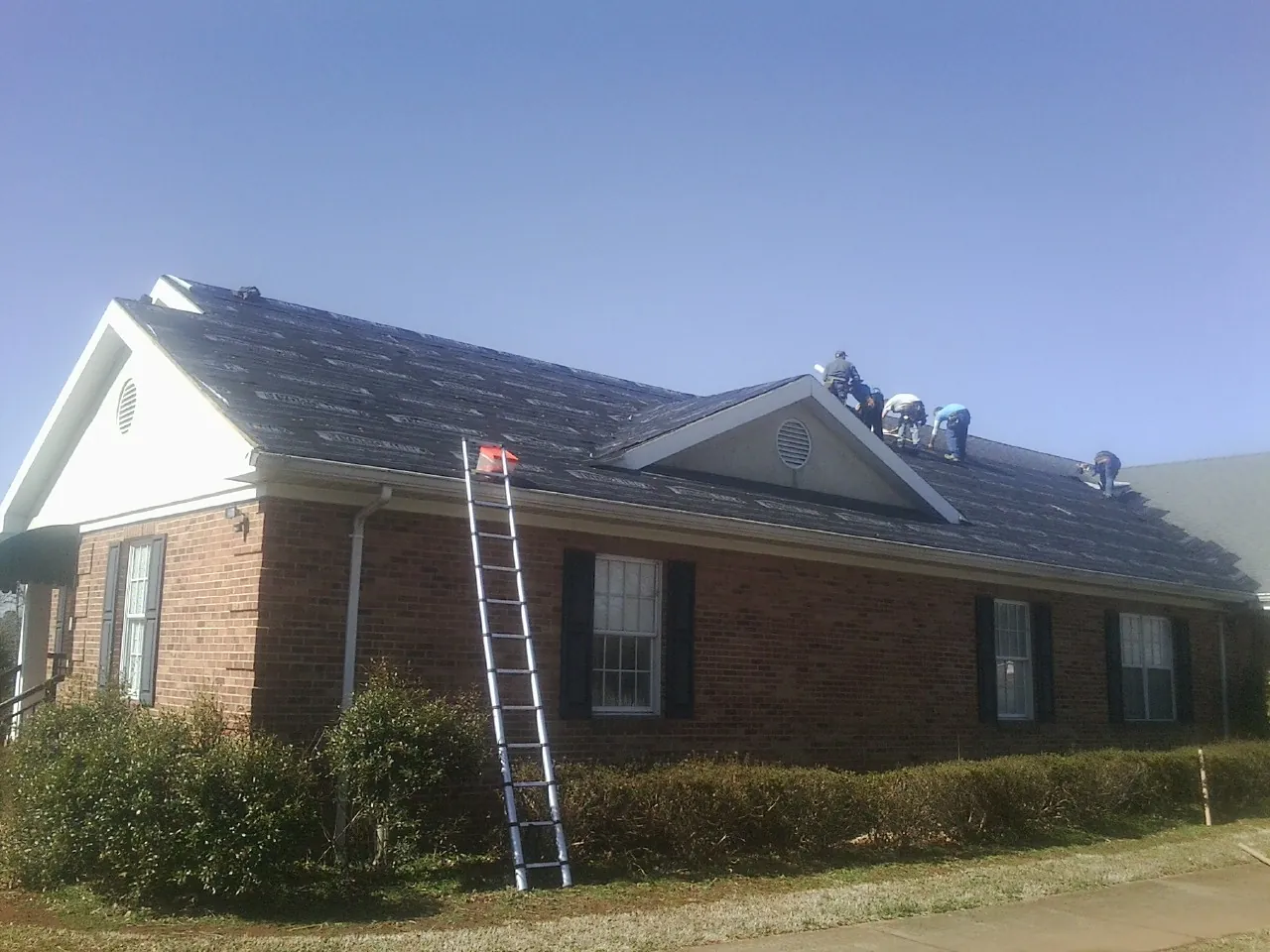 Crew tearing shingles off a brick ranch roof with a ladder set — Emergency Roof Repair work by our crew in Hadley